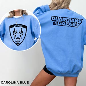 May include: Carolina blue sweatshirt featuring a black Drax graphic on the front and "GUARDIANS OF THE GALAXY" on the back. The Drax graphic is a shield shape with the word "DRAX" above a stylized face. The sweatshirt is a crew neck.