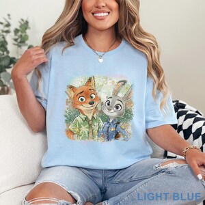 K&ouml;nnte beinhalten: Hellblaues T-Shirt mit einer farbenfrohen Illustration von Nick Wilde und Judy Hopps aus Zoomania. Die Grafik zeigt die Charaktere l&auml;chelnd. Der Text "LIGHT BLUE" befindet sich am unteren Rand des Shirts.