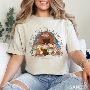 May include: Sand-colored t-shirt featuring a graphic of the Seven Dwarfs from Snow White. The design includes the text "We Dig Dig Dig A-Dig Dig" and "Seven Dwarfs Mine Train" with the year "EST 2014". The dwarves are depicted around a mine entrance.