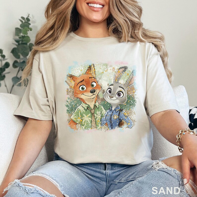 K&ouml;nnte beinhalten: Sandfarbenes T-Shirt mit einer farbenfrohen Illustration von Nick Wilde und Judy Hopps aus Zoomania. Nick tr&auml;gt ein gr&uuml;nes Hemd, und Judy ist in einer Polizeiuniform. Das Wort "SAND" ist unten auf dem T-Shirt aufgedruckt.