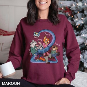 K&ouml;nnte beinhalten: Burgunderfarbenes Sweatshirt mit einer Cartoon-Illustration von Filmfiguren. Das Design umfasst eine blaue Schlange mit der Zahl 2 und den Namen Judy, Nick und Nibbles. Das Wort "MAROON" ist unten aufgedruckt.