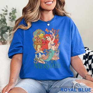 May include: Royal blue t-shirt featuring a graphic of Ariel from The Little Mermaid. The design includes Ariel, Flounder, and floral elements. The text "THE LITTLE MERMAID" is printed in green. The shirt is a solid royal blue color.
