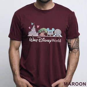 May include: Maroon t-shirt featuring a graphic of Walt Disney World landmarks, including a castle, tree, Mickey Mouse ears, and a sphere. The words "Walt Disney World" are printed in white below the graphic. The shirt is a solid maroon color.
