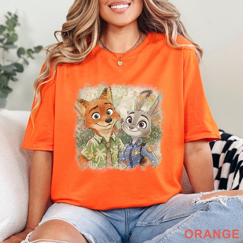 K&ouml;nnte beinhalten: Leuchtend orangefarbenes T-Shirt mit einem Cartoon-Bild von Nick Wilde und Judy Hopps aus Zootopia. Die Grafik zeigt die Charaktere l&auml;chelnd, Nick in einem Hawaiihemd und Judy in einer Polizeiuniform. Das Wort "ORANGE" ist unten aufgedruckt.