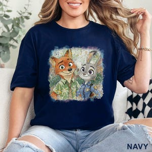 K&ouml;nnte beinhalten: Marineblaues T-Shirt mit einem farbenfrohen Grafikdruck von Nick Wilde und Judy Hopps aus Zootopia. Das Bild zeigt die Charaktere in einem Vintage-Stil. Das Wort "NAVY" ist unten rechts auf dem Shirt aufgedruckt.