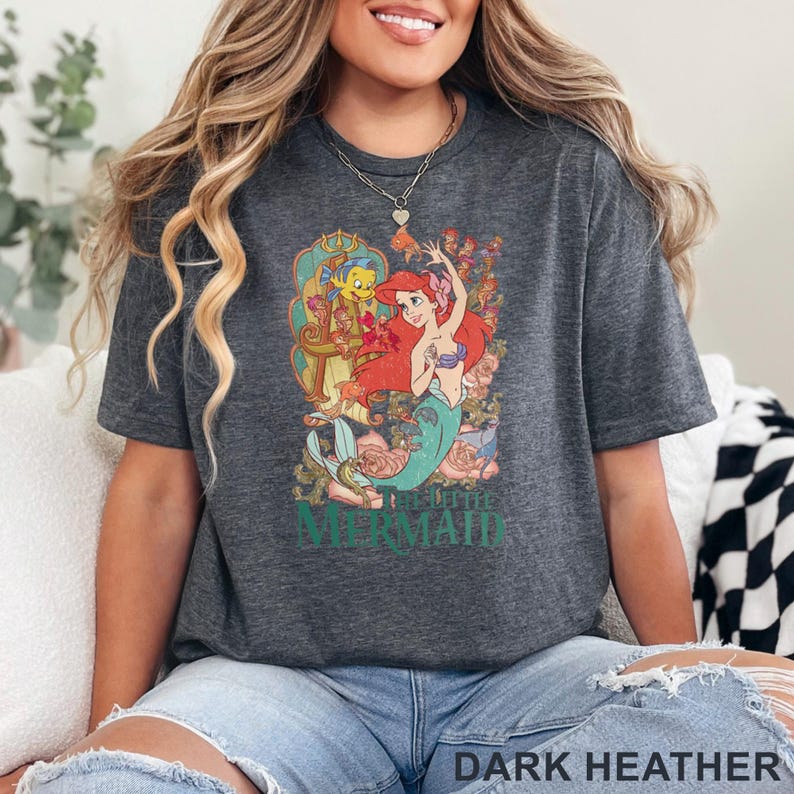 May include: Dark heather gray t-shirt featuring a colorful graphic of Ariel from The Little Mermaid. The design includes Ariel, Flounder, and floral elements. The text "THE LITTLE MERMAID" is printed below the graphic.