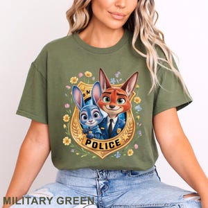 Peut inclure: T-shirt vert militaire avec une image de dessin anim&eacute; d'un renard et d'un lapin en uniforme de police, sur un badge dor&eacute; avec le mot "POLICE". Le motif est entour&eacute; de fleurs.