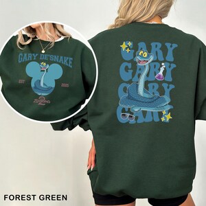 May include: Forest green sweatshirt featuring a cartoon snake character with the text "GARY DE'SNAKE" and "ZOOTOPIA 2". The back of the sweatshirt repeats the name "GARY" with the snake graphic, sunglasses, and stars. The sweatshirt is a crew neck.