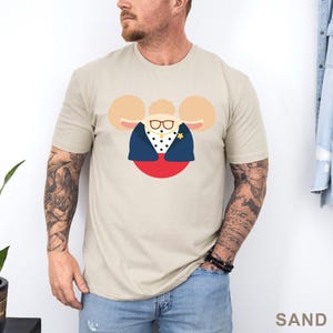 Puede incluir: Camiseta color arena con una ilustraci&oacute;n de dibujos animados de un personaje con orejas grandes, gafas y una chaqueta azul marino sobre una camisa de lunares. El personaje est&aacute; sobre un c&iacute;rculo rojo. La palabra "SAND" est&aacute; impresa abajo a la derecha.