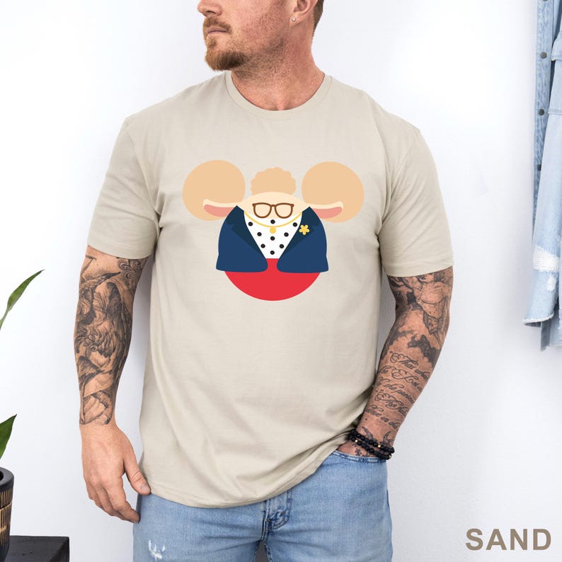Puede incluir: Camiseta color arena con una ilustraci&oacute;n de dibujos animados de un personaje con orejas grandes, gafas y una chaqueta azul marino sobre una camisa de lunares. El personaje est&aacute; sobre un c&iacute;rculo rojo. La palabra "SAND" est&aacute; impresa abajo a la derecha.