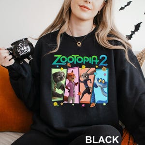 Może przedstawiać: Czarna bluza z napisem "Zootopia 2" i grafiką czterech postaci z filmu animowanego. Słowo "BLACK" jest wydrukowane na dole na biało. Swobodny, wygodny styl ubioru.