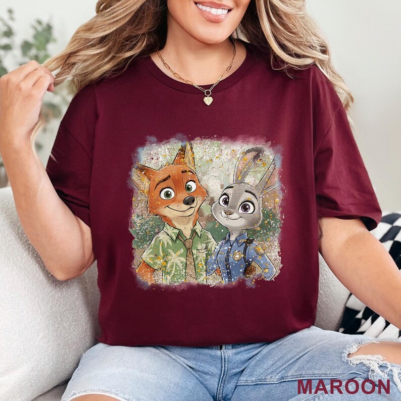 K&ouml;nnte beinhalten: Burgunderfarbenes T-Shirt mit einer Grafik von Nick Wilde und Judy Hopps aus Zootopia. Nick, ein Fuchs, tr&auml;gt ein gr&uuml;nes Hemd, und Judy, ein Hase, ist in einer Polizeiuniform. Das Wort "MAROON" ist unten auf dem Shirt aufgedruckt.