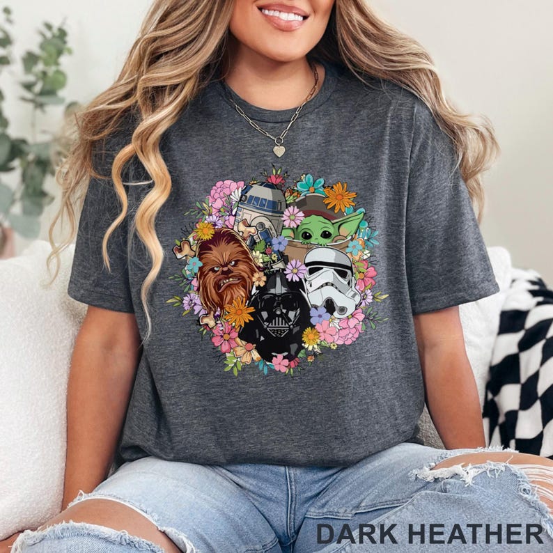 Puede incluir: Camiseta gris jaspeado oscuro con un dise&ntilde;o de corona floral con personajes de Star Wars, incluyendo Darth Vader, Chewbacca y un Stormtrooper. El texto "DARK HEATHER" est&aacute; impreso en la parte inferior.