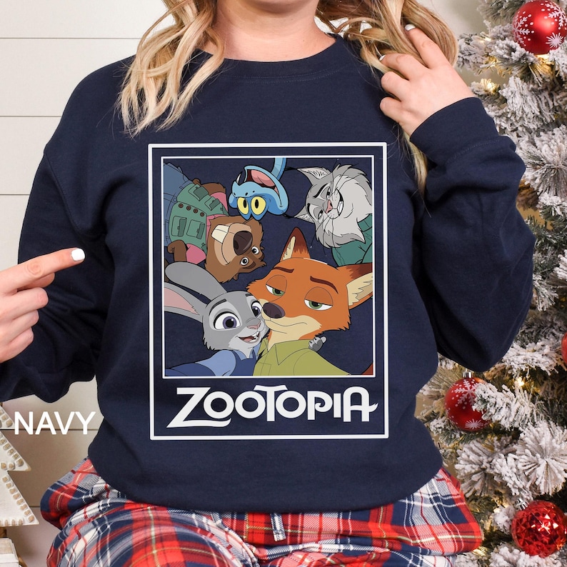 Peut inclure: Sweat-shirt bleu marine avec un motif de "Zootopia". Le design pr&eacute;sente des personnages comme Judy Hopps et Nick Wilde, encadr&eacute;s dans un rectangle blanc. Le mot "ZOOTOPIA" est imprim&eacute; en blanc en dessous, tir&eacute; du film d'animation.