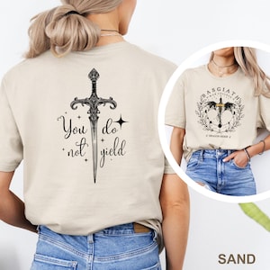 May include: Sand-colored t-shirt featuring a black sword design with the text "You do not yield." A second image shows a similar shirt with a circular design and the text "Basgiath War College Dragon Rider."
