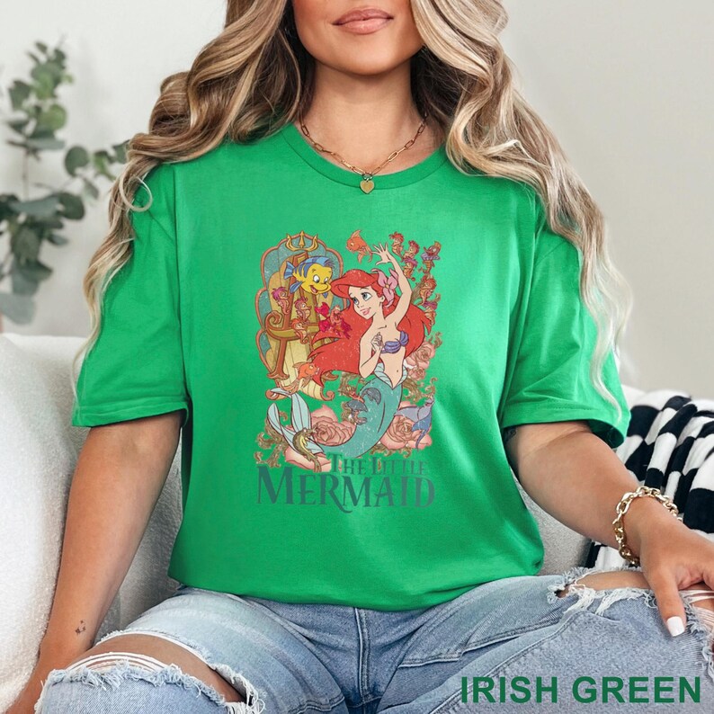 May include: Irish green t-shirt featuring a graphic of Ariel from The Little Mermaid, with Flounder and floral accents. The design is centered on the shirt. The text "THE LITTLE MERMAID" is below the graphic. The shirt is a short-sleeved crew neck.