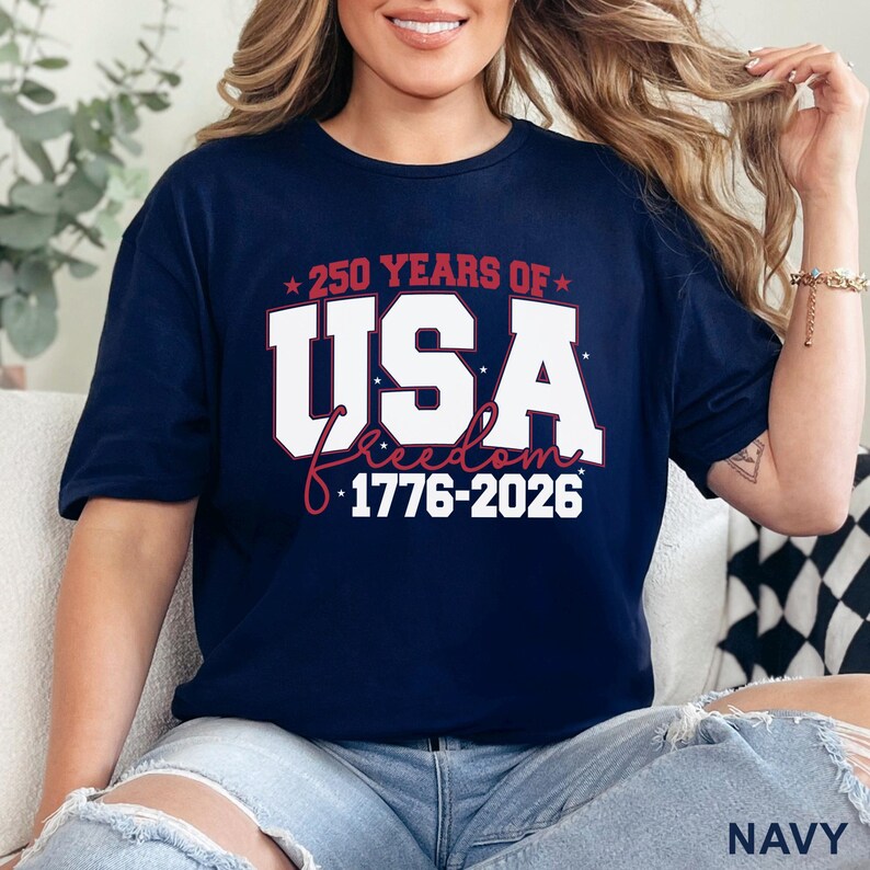 May include: Navy blue t-shirt with the text "250 YEARS OF USA Freedom 1776-2026" in white and red. The shirt is a short-sleeved crew neck style. The word "NAVY" is printed at the bottom right corner of the image.