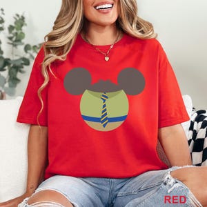 Puede incluir: Camiseta roja con un dise&ntilde;o de personaje de dibujos animados. El dise&ntilde;o incluye orejas marrones, un cuerpo verde y una corbata a rayas azules y amarillas. La palabra "RED" es visible en la esquina inferior derecha.