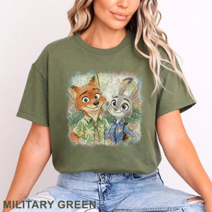 K&ouml;nnte beinhalten: Milit&auml;rgr&uuml;nes T-Shirt mit einem Aufdruck von Nick Wilde und Judy Hopps aus Zootopia. Nick, ein Fuchs, tr&auml;gt ein Hawaiihemd, und Judy, ein Hase, ist in einer Polizeiuniform. Das T-Shirt ist ein bequemer, l&auml;ssiger Stil.