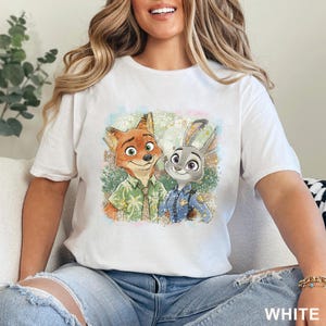K&ouml;nnte beinhalten: Wei&szlig;es T-Shirt mit einer farbenfrohen Illustration von Nick Wilde, dem Fuchs, und Judy Hopps, dem Kaninchen, aus Zoomania. Nick tr&auml;gt ein gr&uuml;nes Hemd und Judy eine blaue Polizeiuniform. Das Wort "WHITE" ist unten aufgedruckt.