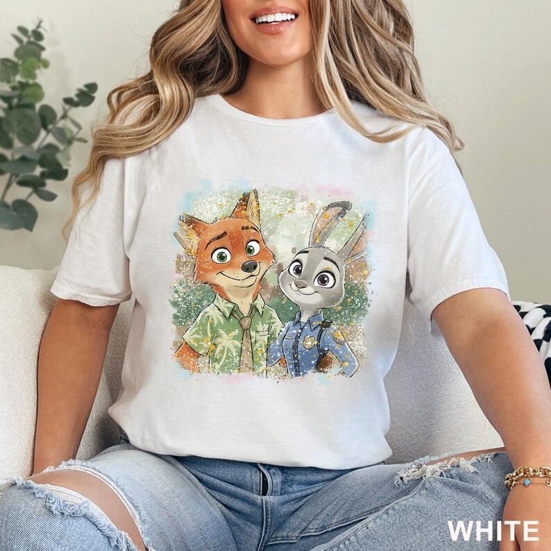 K&ouml;nnte beinhalten: Wei&szlig;es T-Shirt mit einer farbenfrohen Illustration von Nick Wilde, dem Fuchs, und Judy Hopps, dem Kaninchen, aus Zoomania. Nick tr&auml;gt ein gr&uuml;nes Hemd und Judy eine blaue Polizeiuniform. Das Wort "WHITE" ist unten aufgedruckt.
