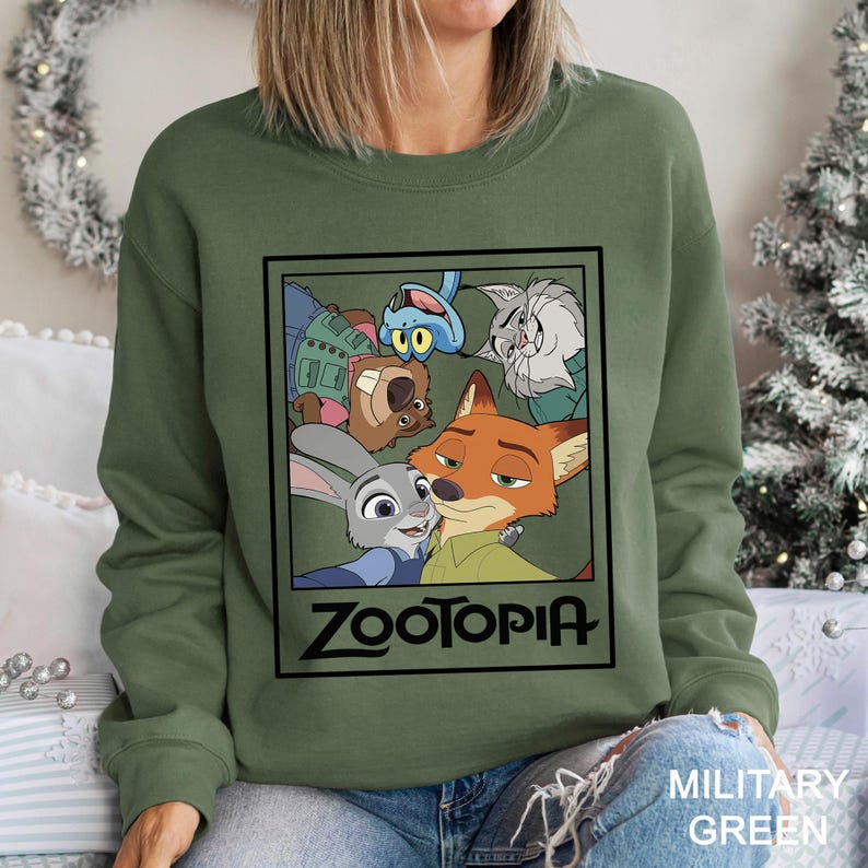 Peut inclure: Sweat-shirt vert militaire avec un motif des personnages du film d'animation Zootopie. Le design comprend Judy Hopps, Nick Wilde et d'autres personnages dans un cadre noir. Le mot "ZOOTOPIA" est imprim&eacute; en dessous.