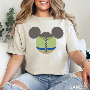 Puede incluir: Camiseta color arena con un gr&aacute;fico de dibujos animados de una cabeza de rat&oacute;n con una camisa verde, corbata a rayas azules y amarillas y orejas grises. La palabra "SAND" est&aacute; impresa en la parte inferior de la camiseta.