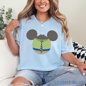 Puede incluir: Camiseta azul claro con un dise&ntilde;o de personaje de dibujos animados. El dise&ntilde;o incluye una camisa verde, una corbata a rayas azules y amarillas y orejas de rat&oacute;n grises. El texto "LIGHT BLUE" est&aacute; impreso en la parte inferior de la camiseta.