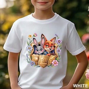 Puede incluir: Camiseta blanca con un dise&ntilde;o colorido de Judy Hopps y Nick Wilde de Zootopia, enmarcado por flores y una placa de polic&iacute;a. El dise&ntilde;o incluye el texto "Zootopia Police".