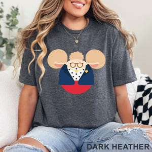 Puede incluir: Camiseta gris oscuro con un dise&ntilde;o de personaje de dibujos animados. El dise&ntilde;o incluye un personaje con gafas, una chaqueta azul y un broche de flor amarilla. El texto "DARK HEATHER" est&aacute; en la parte inferior de la imagen.