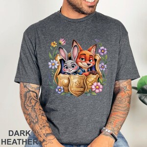 Puede incluir: Camiseta gris jaspeado oscuro con un gr&aacute;fico colorido de Judy Hopps y Nick Wilde de Zootopia, enmarcado por flores y placas de polic&iacute;a. El texto "Zootopia Police" es visible en la placa. La camiseta es de material suave.