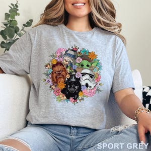 Puede incluir: Camiseta gris deportiva con un dise&ntilde;o de corona floral con personajes de Star Wars: Chewbacca, Darth Vader, un Stormtrooper, R2-D2 y Grogu. La camiseta tiene mangas cortas y cuello redondo. El texto "SPORT GREY" est&aacute; impreso en la parte inferior.