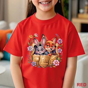 Puede incluir: Camiseta roja con un gr&aacute;fico colorido de Judy Hopps y Nick Wilde de Zootopia, enmarcado por flores y una placa de polic&iacute;a. La palabra "RED" est&aacute; impresa en la esquina inferior derecha de la camiseta.