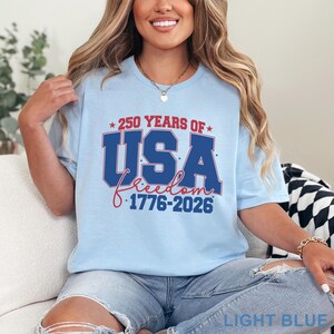May include: Light blue t-shirt with the text "250 YEARS OF USA Freedom 1776-2026" in red and blue. The shirt is a crew neck style and is made of a soft material. The shirt is a light blue color.