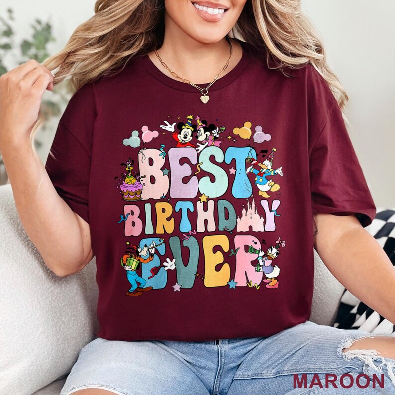 May include: Maroon t-shirt with the text "BEST BIRTHDAY EVER" in colorful bubble letters. The design includes cartoon characters like Mickey and Minnie Mouse, Donald Duck, and Goofy, with a birthday cake and castle.