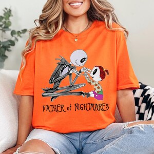 Puede incluir: Camiseta naranja brillante con un dise&ntilde;o de Jack Skellington y un ni&ntilde;o. El texto "FATHER OF NIGHTMARES" est&aacute; impreso debajo. La camiseta est&aacute; hecha de un material suave. Una camiseta divertida e informal.
