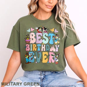 May include: Military green t-shirt with the text "BEST BIRTHDAY EVER" in colorful bubble letters. The design includes cartoon characters and pastel-colored shapes. The shirt is a crew neck and has short sleeves.