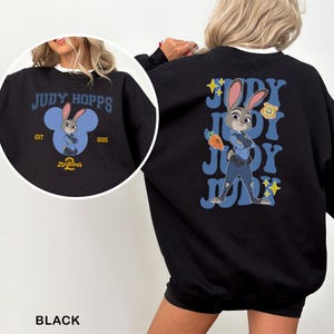 May include: Black sweatshirt featuring Judy Hopps from Zootopia 2. The front design includes the text "JUDY HOPPS" above a cartoon image of Judy Hopps inside a blue Mickey Mouse-shaped design. The back features Judy Hopps with the text "JUDY" repeated.