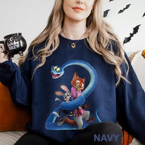 Op de afbeelding: Marineblauwe sweatshirt met een cartoonafbeelding van een blauwe slang die zich om een vos en een konijn wikkelt. De vos draagt een roze shirt, het konijn een pak. Het woord "NAVY" staat onderaan gedrukt. Halloween thema.