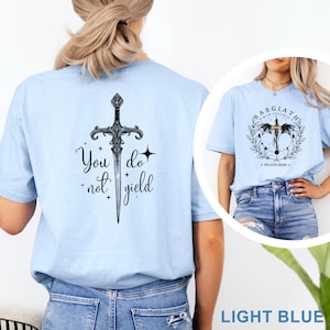 May include: Light blue t-shirt featuring a detailed black sword graphic with the words "You do not yield." A smaller image shows a similar shirt with a circular design and the text "Basgiath War College" and "Dragon Rider V."