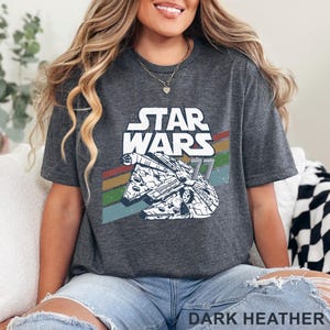 May include: Dark heather gray t-shirt featuring the Star Wars logo and a graphic of the Millennium Falcon. The design includes a retro stripe in red, orange, green, and blue. The text "DARK HEATHER" is at the bottom.