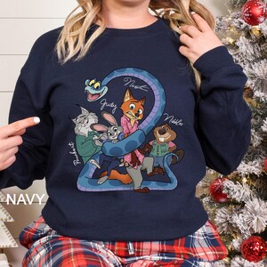 K&ouml;nnte beinhalten: Marineblaues Sweatshirt mit einem farbenfrohen Cartoon-Motiv von Zootopia-Charakteren, darunter Judy Hopps und Nick Wilde. Das Wort "NAVY" ist unten links aufgedruckt.