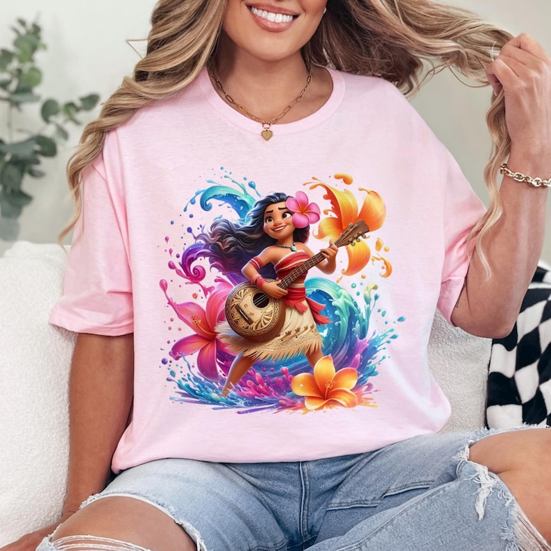 May include: A light pink t-shirt featuring a colorful illustration of a cartoon character playing a ukulele. The character is surrounded by flowers and water splashes in shades of blue, purple, and orange. The shirt is a crew neck style.