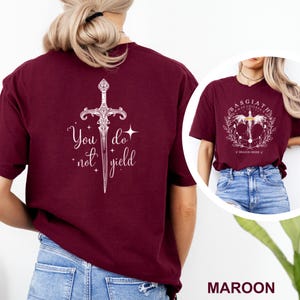 May include: Maroon t-shirt with a white graphic of a sword and the text "You do not yield." A second image shows a similar shirt with a circular design and the text "Basgiath War College."