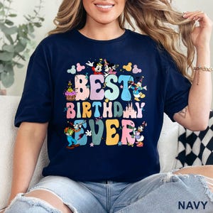 May include: Navy blue t-shirt with the text "BEST BIRTHDAY EVER" in colorful bubble letters. The design includes cartoon characters like Mickey and Minnie Mouse, Donald Duck, and Goofy, with stars and a castle.