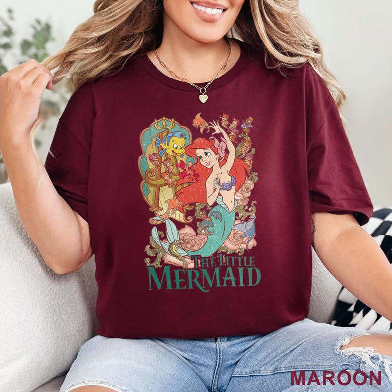May include: Maroon t-shirt featuring a colorful graphic of Ariel and Flounder from The Little Mermaid. The design includes the text "THE LITTLE MERMAID" in green. The shirt is a solid maroon color.