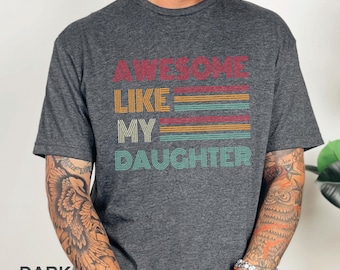 May include: Dark heather gray t-shirt with the text "AWESOME LIKE MY DAUGHTER" in a retro design. The text is in red, yellow, teal, and maroon. The shirt is a crew neck and has short sleeves.