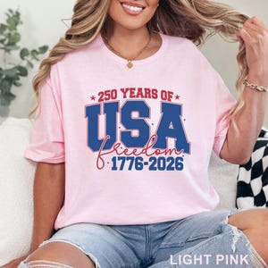 May include: Light pink t-shirt with the text "250 YEARS OF USA Freedom 1776-2026" in red, white, and blue. The shirt is a crew neck and has short sleeves. The words "LIGHT PINK" are at the bottom of the image.