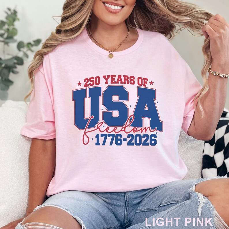 May include: Light pink t-shirt with the text "250 YEARS OF USA Freedom 1776-2026" in red, white, and blue. The shirt is a crew neck and has short sleeves. The words "LIGHT PINK" are at the bottom of the image.