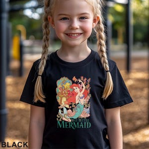 May include: Black t-shirt featuring a colorful graphic of Ariel from The Little Mermaid with the text "THE LITTLE MERMAID" in green. The shirt is worn by a young girl with blonde braids.